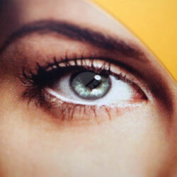 Nahaufnahme eines menschlichen Auges mit grünem Iris und Wimpern vor einem gelben Hintergrund.