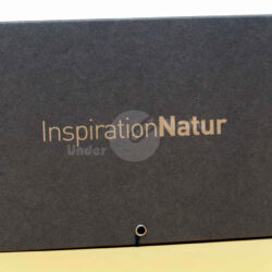 Schwarzes Notizbuch mit goldenem Schriftzug "InspirationNatur" und Klemmmechanismus auf einem hellen Hintergrund.