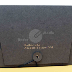 Graue Papiermappe mit goldener Aufschrift "Katholische Akademie Stapelfeld", verschlossen mit einem blauen Band und einer Öse.