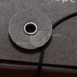 Nahaufnahme einer schwarzen Pappscheibe mit einer zentralen Metallöse, die an einem schwarzen Band befestigt ist.