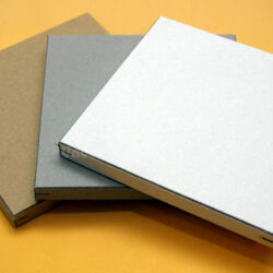 Drei quadratische Pappverpackungen in braun, grau und weiß auf einem gelben Hintergrund.