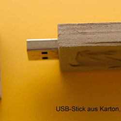Ein kartonbasierter USB-Stick liegt auf einem gelben Hintergrund, mit einem grauen Schutzdeckel daneben.