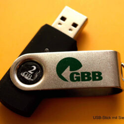 Nahaufnahme eines USB-Sticks mit Siebdruck des Logos "GBB" auf silbernem und schwarzem Gehäuse.
