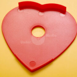 Eine rote, herzförmige Kunststoffscheibe mit einem zentralen Loch und drei Befestigungsstellen, auf einem gelben Hintergrund.